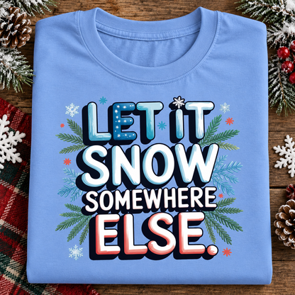 A folded carolina blue t-shirt with a festive graphic reading 'LET IT SNOW SOMEWHERE ELSE.' surrounded by snowflakes, pine branches, and small red stars, displayed on a rustic wooden surface with holiday decorations.