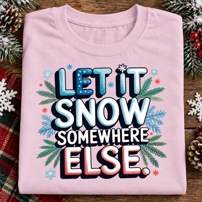 A folded light pink t-shirt with a festive graphic that reads 'LET IT SNOW SOMEWHERE ELSE.' in blue, white, and red lettering, surrounded by green pine branches and snowflake decorations, displayed on a rustic wooden background with holiday pinecones and a plaid blanket.