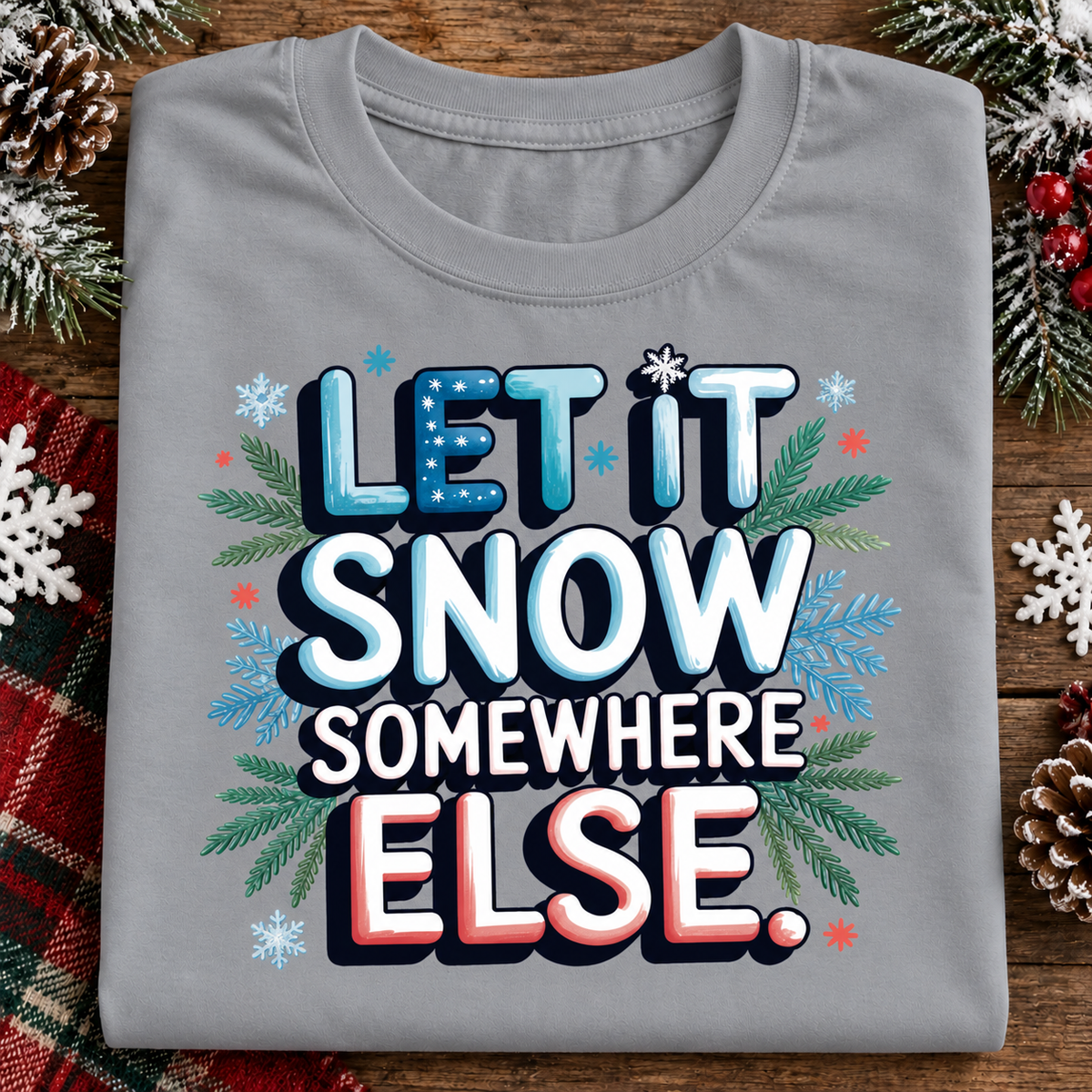 A folded sport gray t-shirt with a festive graphic reading 'LET IT SNOW SOMEWHERE ELSE.' in blue, white, and red lettering, surrounded by snowflakes, pine branches, and red berries on a rustic wooden background.