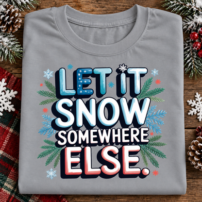 A folded sport gray t-shirt with a festive graphic reading 'LET IT SNOW SOMEWHERE ELSE.' in blue, white, and red lettering, surrounded by snowflakes, pine branches, and red berries on a rustic wooden background.