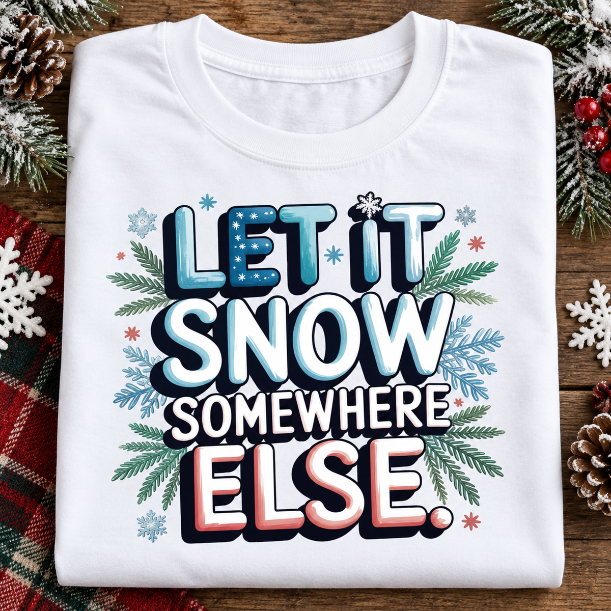 A folded white graphic t-shirt with the text 'LET IT SNOW SOMEWHERE ELSE.' in stylized blue, white, and pink lettering, surrounded by green pine branches and snowflakes, displayed on a rustic wooden background with holiday decorations.