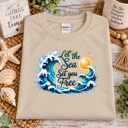A sand colored gildan softstyle t-shirt with a graphic design with the words "Let the Seas set you free" surrounded by ocean waves and a sun