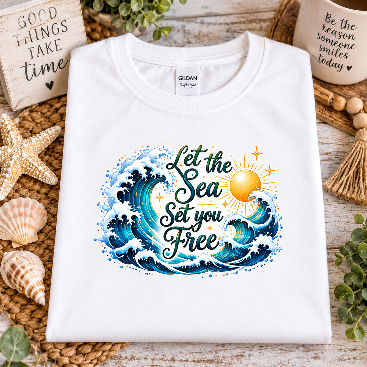 A white gildan softstyle t-shirt with a graphic design with the words "Let the Seas set you free" surrounded by ocean waves and a sun