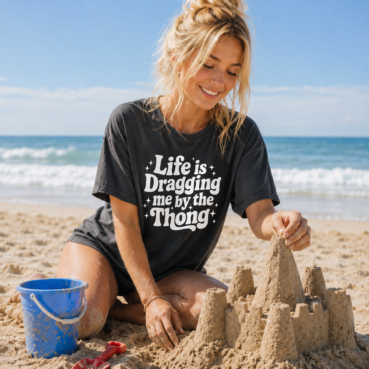 Woman building a sandcastle on the beach wearing a black t-shirt with text.