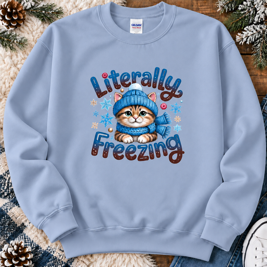 A light blue crewneck sweatshirt featuring a graphic of a cute tabby kitten wearing a blue knit hat and scarf, surrounded by the text 'Literally Freezing' and small snowflakes. The sweatshirt is laid flat on a rustic wood background with faux fur, pinecones, and jeans.