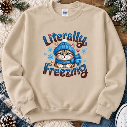 A sand crewneck sweatshirt featuring a graphic of a cute orange tabby kitten wearing a blue knit hat and scarf, surrounded by the text 'Literally Freezing' in a stylized font. The sweatshirt is laid flat on a rustic wooden surface decorated with pinecones, a white blanket, and blue jeans.