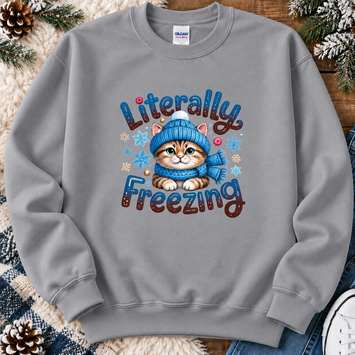 A sport gray crewneck sweatshirt featuring a graphic of a cute orange tabby kitten wearing a blue knit hat and matching blue scarf, surrounded by the text 'Literally Freezing' and small snowflakes. The sweatshirt is laid flat on a rustic wood background with holiday pine branches, a white faux fur blanket, and a blue plaid blanket.