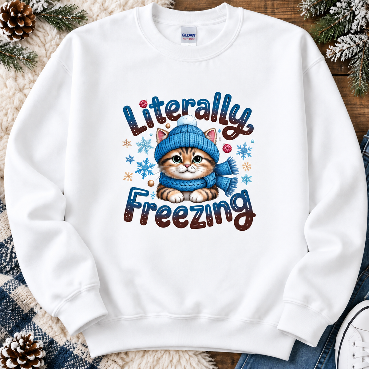 A white crewneck sweatshirt featuring a graphic of a cute orange tabby kitten wearing a blue knit hat and scarf, surrounded by the text 'Literally Freezing' and snowflakes. The sweatshirt is displayed in a flat-lay arrangement with pinecones, a plaid blanket, and denim jeans.