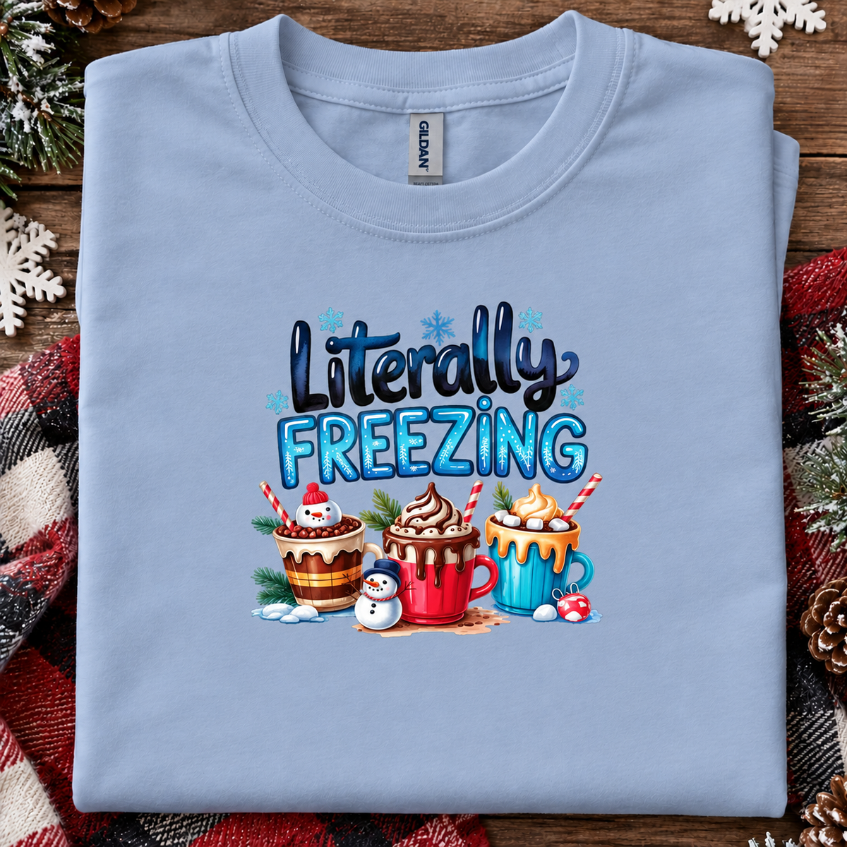 A light blue folded t-shirt with a 'Literally FREEZING' holiday graphic, displayed on a rustic wooden background with a red plaid blanket and pine decorations.