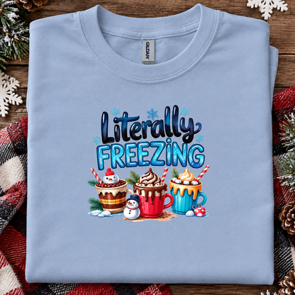 A light blue folded t-shirt with a 'Literally FREEZING' holiday graphic, displayed on a rustic wooden background with a red plaid blanket and pine decorations.