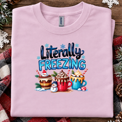 A folded light pink Gildan t-shirt with a festive graphic that reads 'Literally FREEZING' above three hot cocoa drinks and a snowman, styled on a rustic wooden background with holiday decorations.