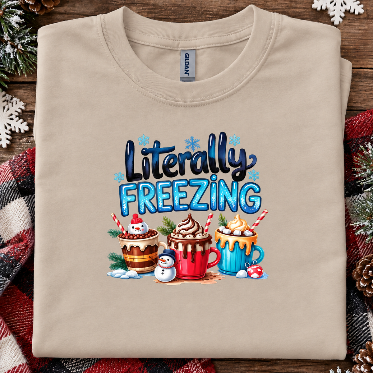 A sand colored Gildan t-shirt with a 'Literally FREEZING' graphic print featuring three hot cocoa mugs and a snowman, laid flat on a rustic wooden background with a plaid blanket and pine decorations.