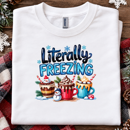 A folded white Gildan t-shirt with a colorful graphic that reads 'Literally FREEZING' above three hot cocoa drinks and a snowman, displayed on a rustic wooden background with holiday decorations.