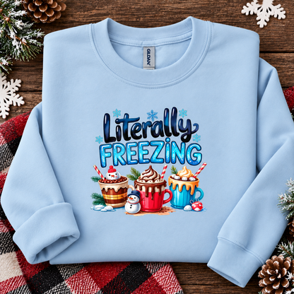 A light blue crewneck sweatshirt with a festive graphic reading 'Literally FREEZING' above three hot cocoa mugs and a snowman, styled on a rustic wooden background with pine branches, snowflakes, and a plaid blanket.