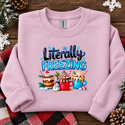 A light pink crewneck sweatshirt with a festive graphic that reads 'Literally FREEZING' above three hot cocoa mugs and snowman decorations, laid flat on a rustic wooden background with holiday decorations.