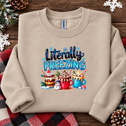 A sand colored crewneck sweatshirt laid flat on a rustic wooden surface, decorated with pine branches, snowflakes, and a plaid blanket. The sweatshirt features a colorful graphic that reads 'Literally FREEZING' above three hot cocoa mugs and snowman illustrations.