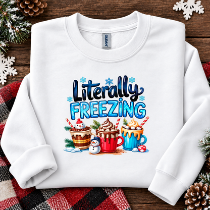 A white long-sleeve crewneck sweatshirt laid flat on a rustic wooden surface with holiday decorations. The front features a colorful graphic that reads 'Literally FREEZING' above three hot cocoa cups, a snowman, and small snowflakes.