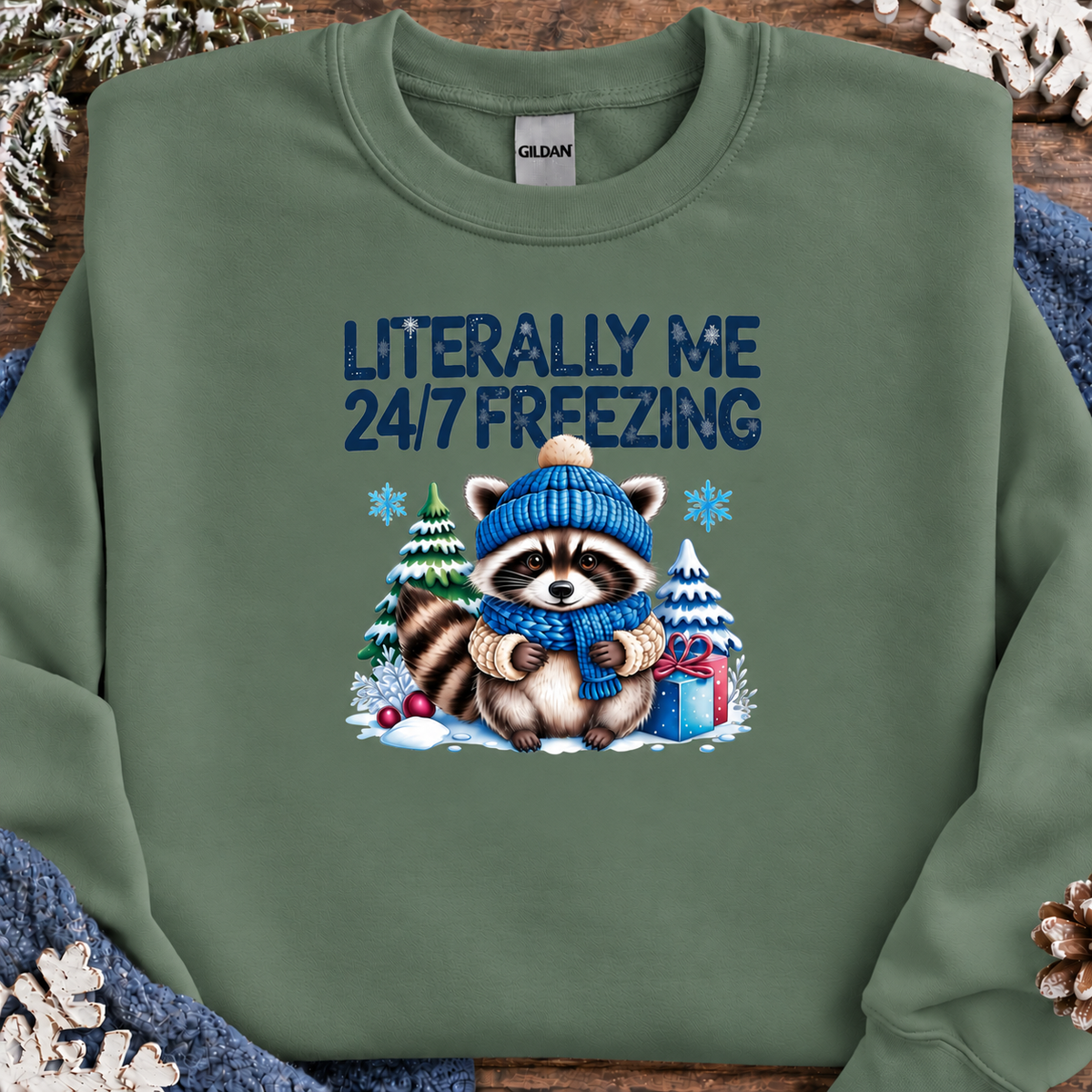 A military green crewneck sweatshirt by Gildan, featuring a winter-themed graphic of a raccoon in a blue knit hat and scarf surrounded by snow, pine trees, and a gift box, with the text 'LITERALLY ME 24/7 FREEZING' printed above. The sweatshirt is styled flat on a rustic wooden background with holiday decorations.