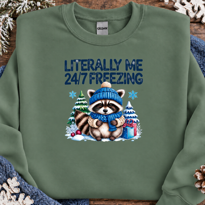 A military green crewneck sweatshirt by Gildan, featuring a winter-themed graphic of a raccoon in a blue knit hat and scarf surrounded by snow, pine trees, and a gift box, with the text 'LITERALLY ME 24/7 FREEZING' printed above. The sweatshirt is styled flat on a rustic wooden background with holiday decorations.