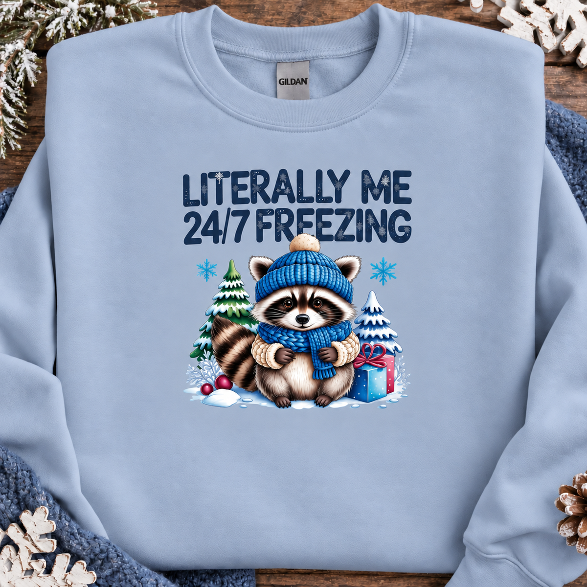 A light blue crewneck sweatshirt by Gildan, featuring a winter-themed graphic of a raccoon in a blue beanie and scarf surrounded by snow, pine trees, and a gift box, with the text 'LITERALLY ME 24/7 FREEZING'. The sweatshirt is styled flat on a rustic wooden background with holiday decorations.