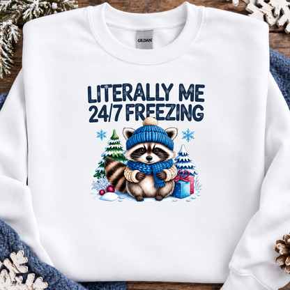 A white Gildan crewneck sweatshirt laid flat on a rustic wooden surface with winter-themed props. The sweatshirt features a graphic of a raccoon wearing a blue knit hat and matching blue scarf, surrounded by snow, Christmas trees, and a gift box, with the text 'LITERALLY ME 24/7 FREEZING' printed above.
