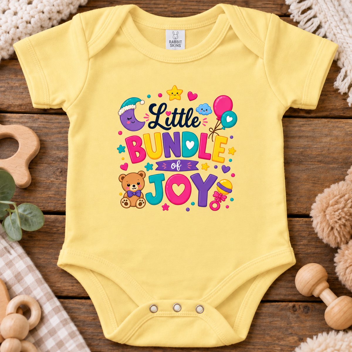 A butter yellow short-sleeve baby bodysuit laid flat on a rustic wooden surface, featuring a colorful 'Little Bundle of Joy' graphic with a teddy bear, balloons, stars, and hearts. 