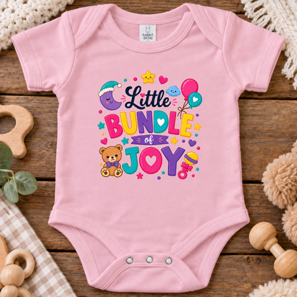 A pink short-sleeve baby bodysuit laid flat on a rustic wooden surface, featuring a colorful graphic that reads 'Little BUNDLE of JOY' with a teddy bear, balloons, stars, and hearts. 