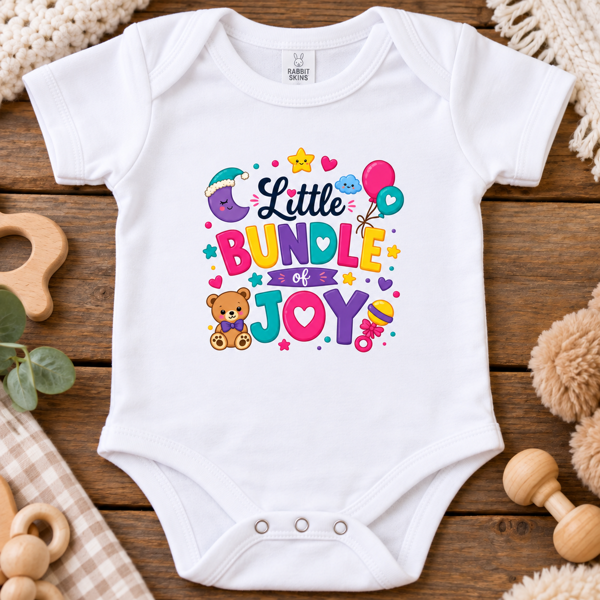 A white short-sleeve baby bodysuit laid flat on a rustic wooden surface, featuring a colorful 'Little Bundle of Joy' graphic with a teddy bear, balloons, stars, and hearts. 