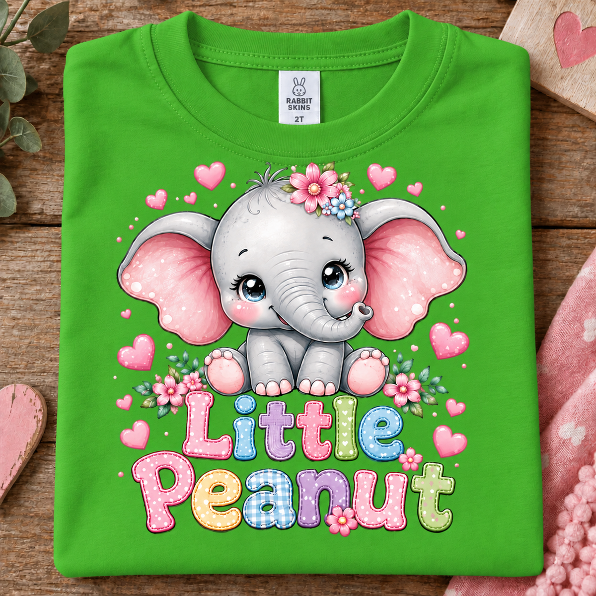 A folded apple green t-shirt with a cartoon graphic of a cute gray elephant with pink ears, surrounded by pink hearts and flowers, with the text 'Little Peanut' in colorful letters. The shirt is displayed on a rustic wooden surface with decorative props.