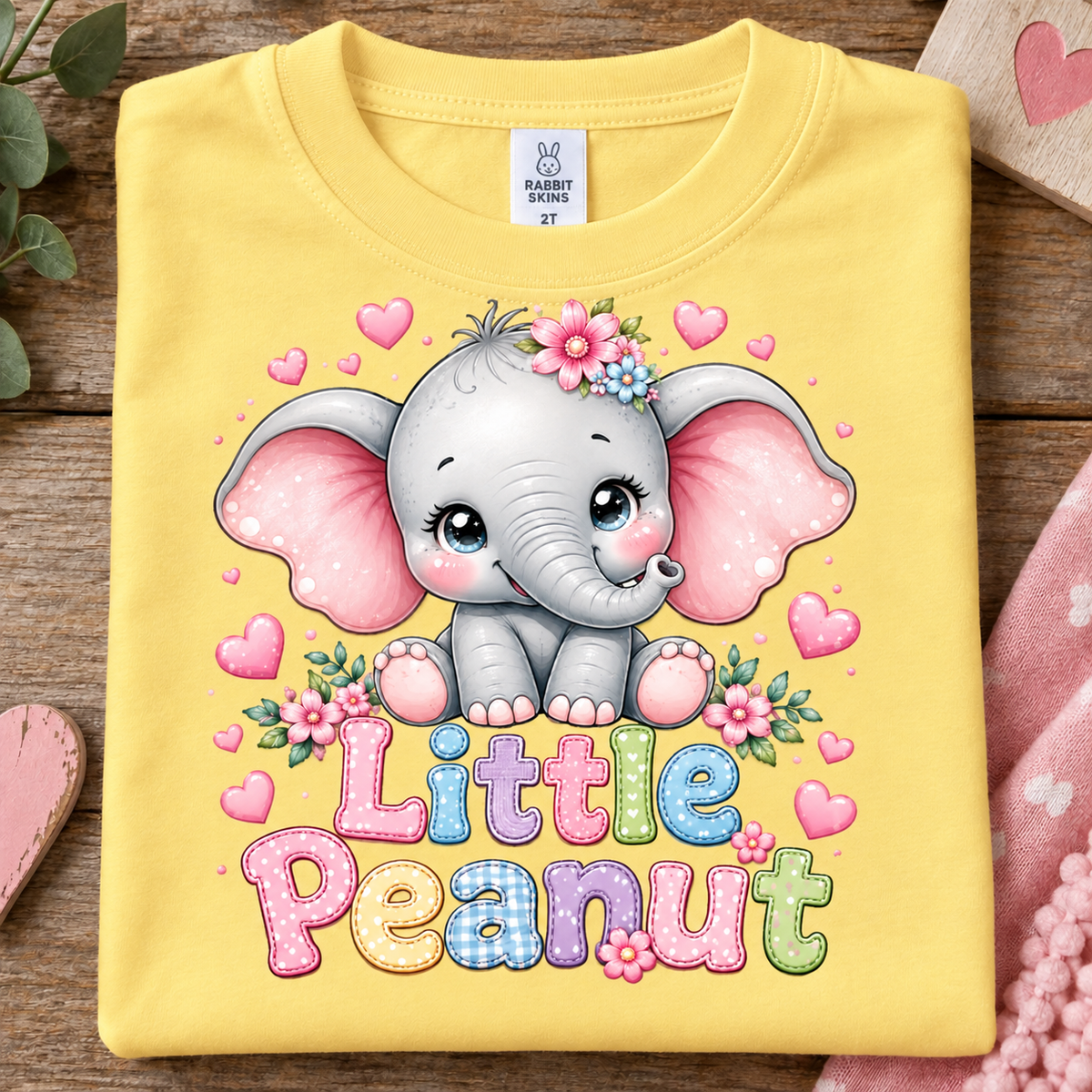 A folded butter colored t-shirt with a colorful cartoon graphic of a baby elephant and the text 'Little Peanut' on a rustic wooden background.