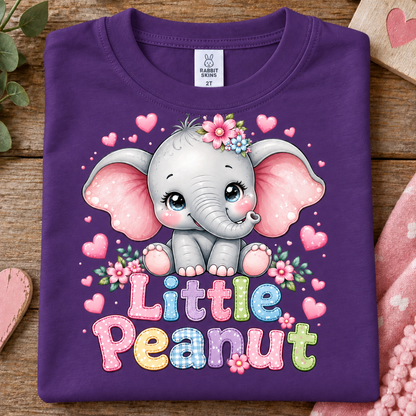 A folded purple t-shirt on a rustic wooden surface, featuring a cute cartoon baby elephant with pink ears, a flower crown, and the text 'Little Peanut' in colorful letters.