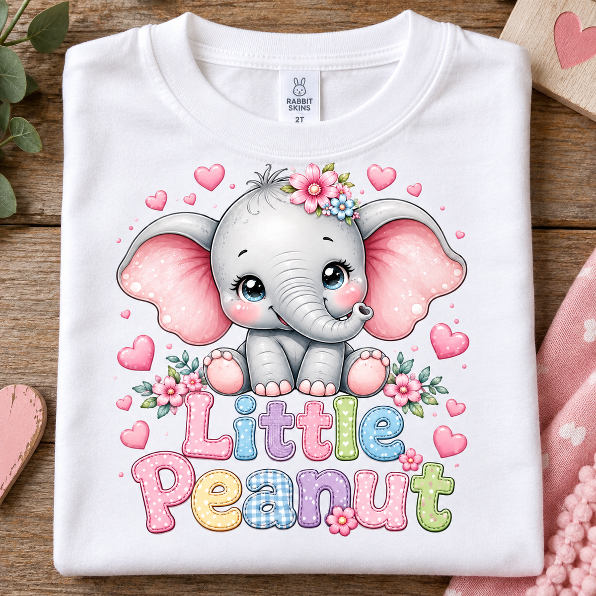 A folded white baby t-shirt with a cute cartoon elephant graphic and the text 'Little Peanut' in colorful letters, displayed on a rustic wooden background with pink decorative props.