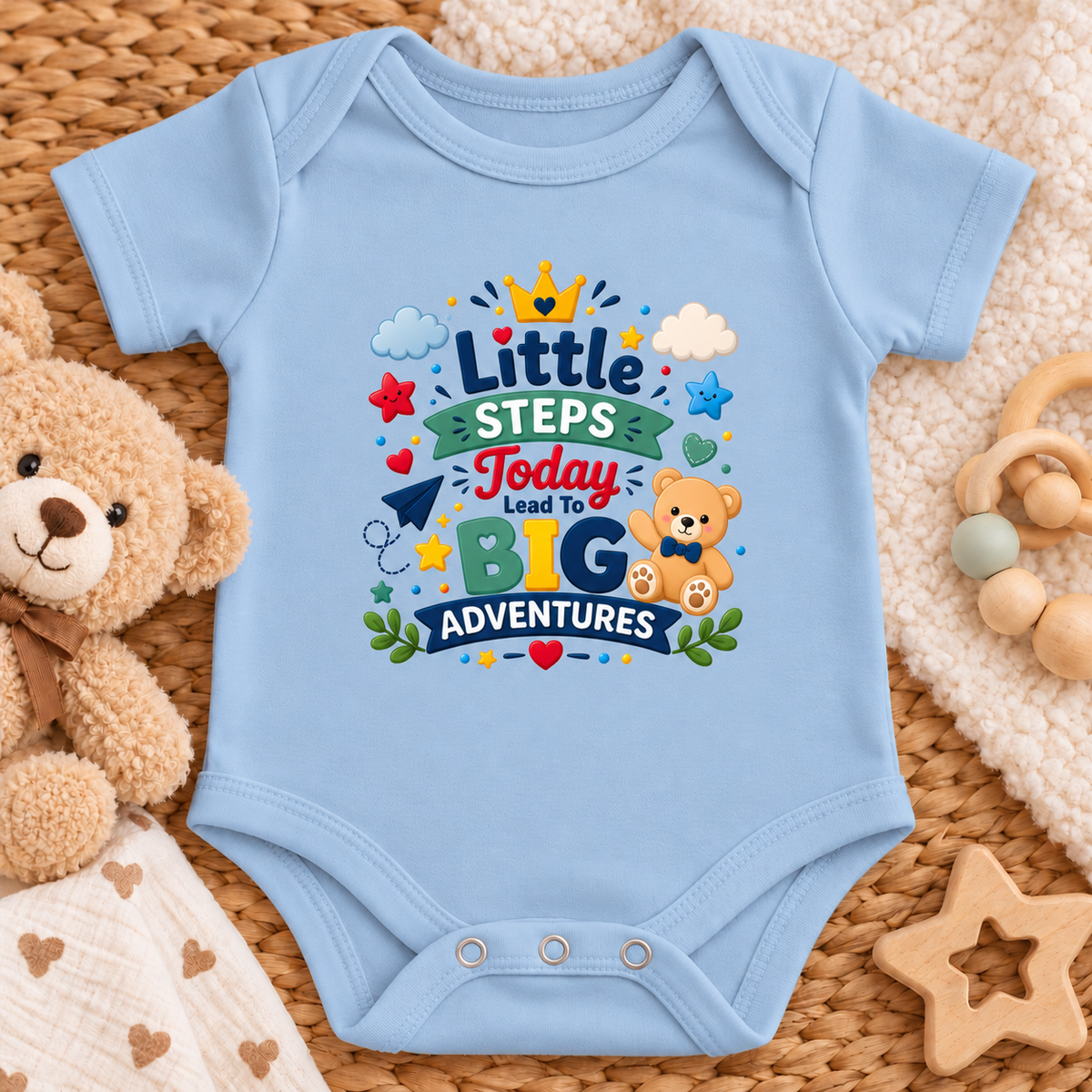 A light blue baby bodysuit laid flat on a woven mat, surrounded by a teddy bear, wooden toy, and soft blanket. The bodysuit features a colorful graphic that reads 'Little Steps Today Lead To BIG Adventures' with cartoon stars, clouds, a crown, and a teddy bear.