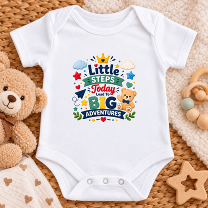 A white short-sleeve baby bodysuit laid flat on a woven mat, surrounded by a teddy bear, wooden toys, and a cream blanket. The bodysuit features a colorful graphic on the chest that reads 'Little Steps Today Lead To BIG Adventures' with stars, clouds, a crown, and a teddy bear.