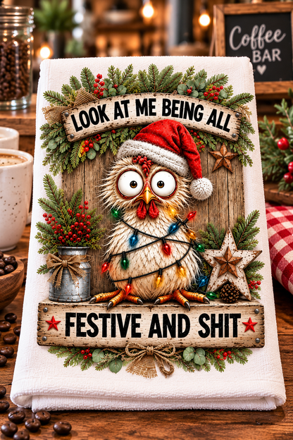 A white kitchen towel featuring a festive Christmas owl design with a Santa hat, colorful holiday lights, pine garlands, and humorous text reading 'LOOK AT ME BEING ALL' and 'FESTIVE AND SHIT', displayed on a rustic wooden table with coffee and holiday decor in the background.
