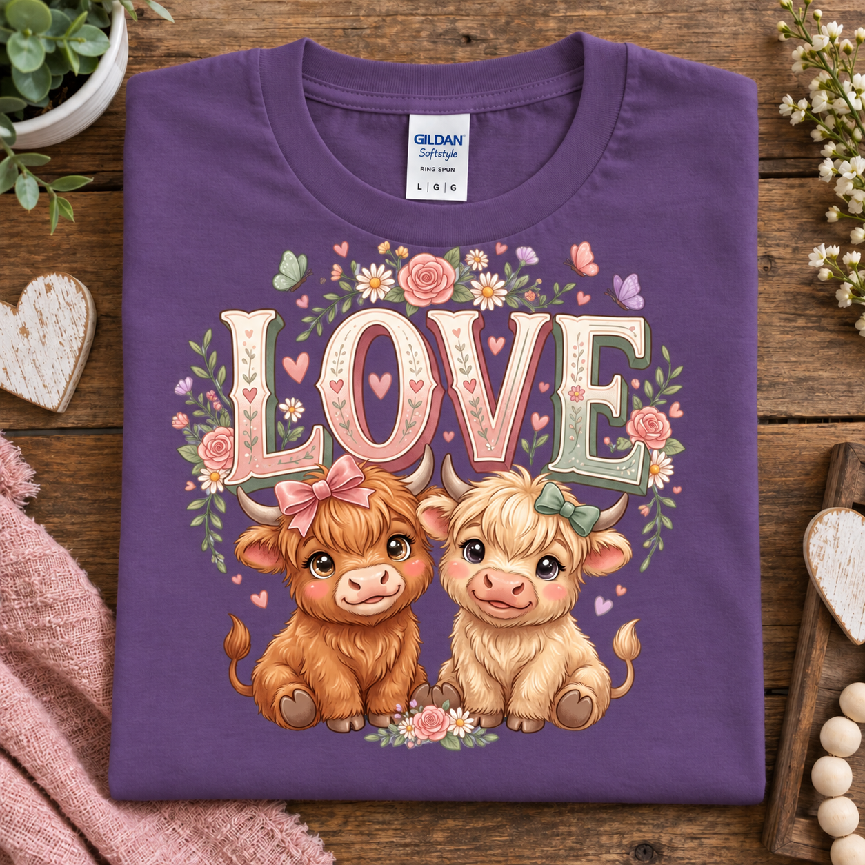 A folded purple Gildan Straight cut T-shirt with a colorful graphic of two cute Highland cows under the word 'LOVE' surrounded by flowers, displayed on a rustic wooden surface with decorative props.