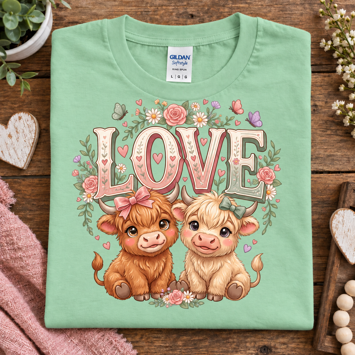 A folded mint green Gildan Blue Work t-shirt with a cute graphic of two Scottish Highland cows under the word 'LOVE' decorated with flowers and hearts, displayed on a rustic wooden table with cozy decorative props.
