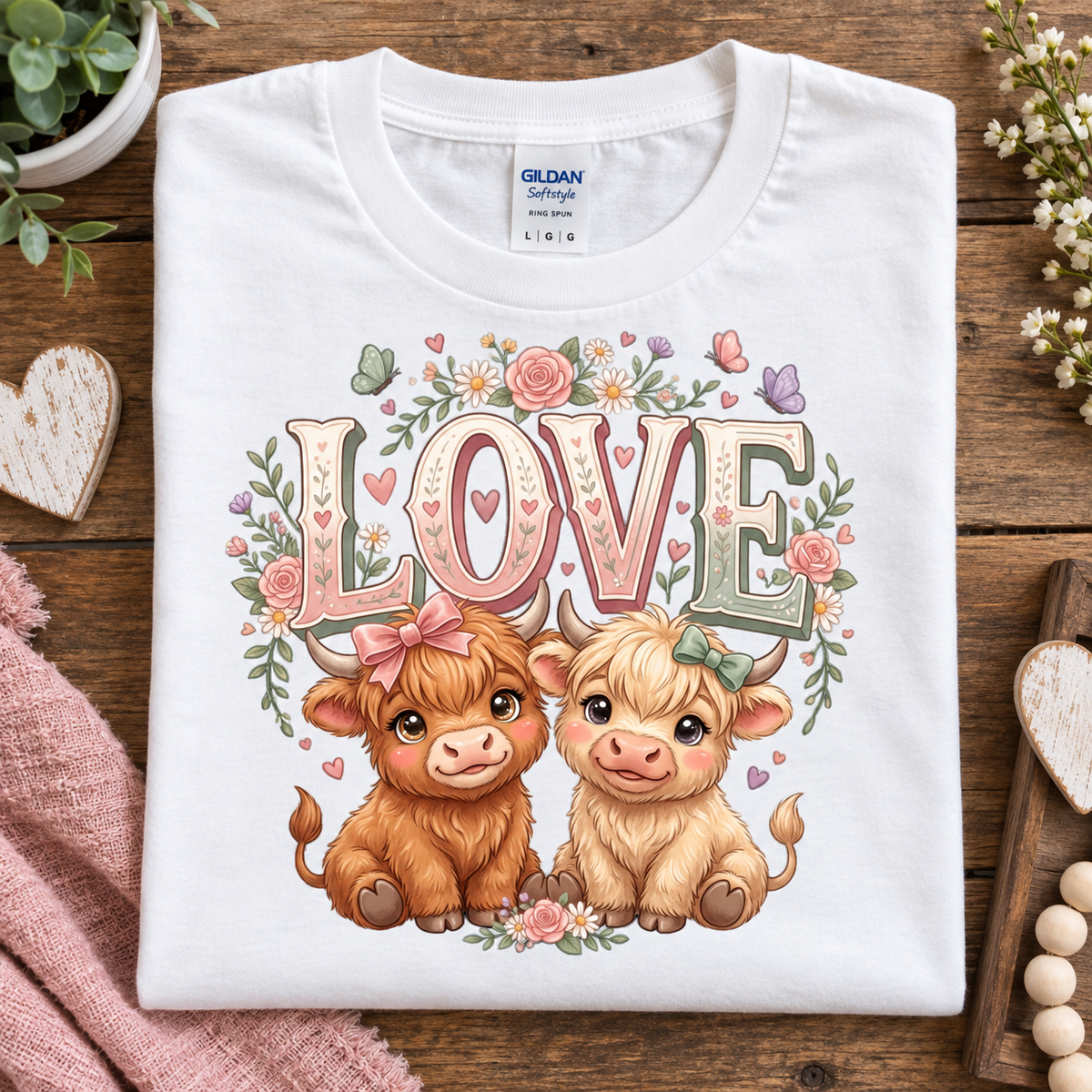 A folded white Gildan Softstyle T-shirt with a colorful 'LOVE' graphic featuring two cute cartoon cows, displayed on a rustic wooden surface with decorative props.