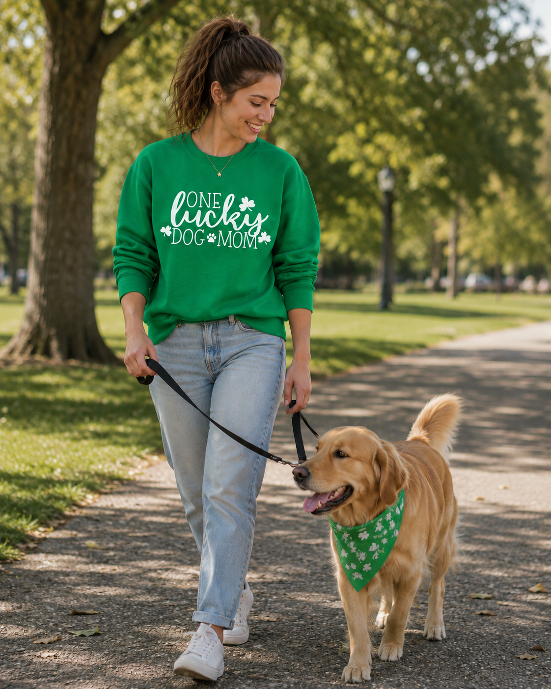 Woman walking a dog on a leash in a park wearing a green sweatshirt with One Lucky Dog Mom text.