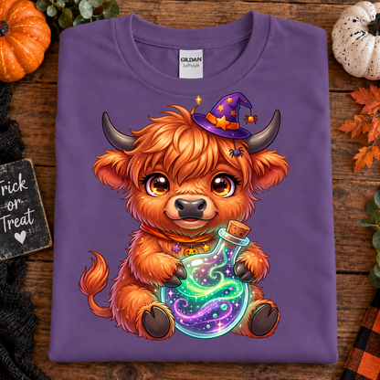 A heather purple Gildan t-shirt laid flat on a rustic wooden surface, featuring a cartoon illustration of a fluffy orange Highland cow wearing a purple witch hat and holding a glowing multicolor potion bottle. The shirt is styled with autumn props including a small pumpkin, a white ornament, fall leaves, and a 'Trick or Treat' sign.