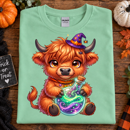 A folded mint green Gildan t-shirt with a colorful cartoon graphic of a fluffy orange Highland cow wearing a purple witch hat and holding a glowing potion bottle. The shirt is displayed on a rustic wooden background decorated with autumn leaves, a small pumpkin, and a 'Trick or Treat' sign.