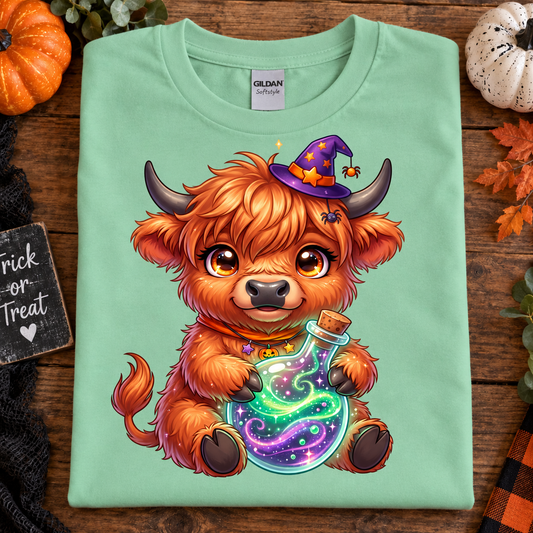 A folded mint green Gildan t-shirt with a colorful cartoon graphic of a fluffy orange Highland cow wearing a purple witch hat and holding a glowing potion bottle. The shirt is displayed on a rustic wooden background decorated with autumn leaves, a small pumpkin, and a 'Trick or Treat' sign.