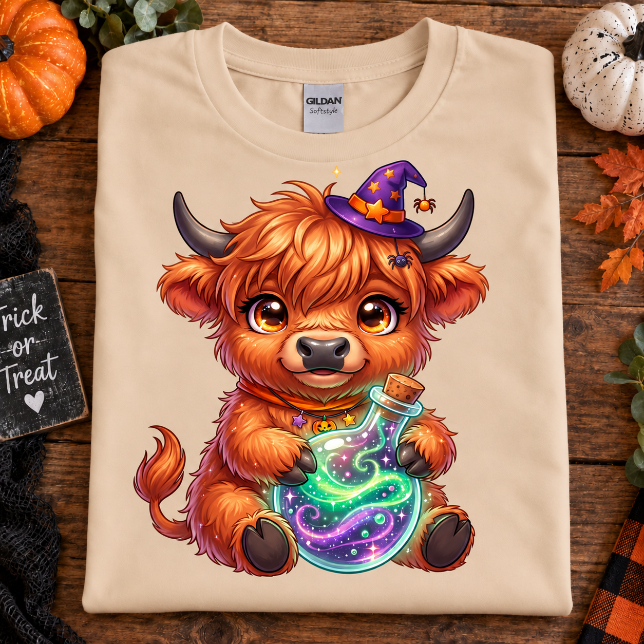 A folded natural Gildan t-shirt with a colorful cartoon graphic of a fluffy orange Highland cow wearing a purple witch hat and holding a glowing potion bottle, styled on a rustic wooden background with autumn props.