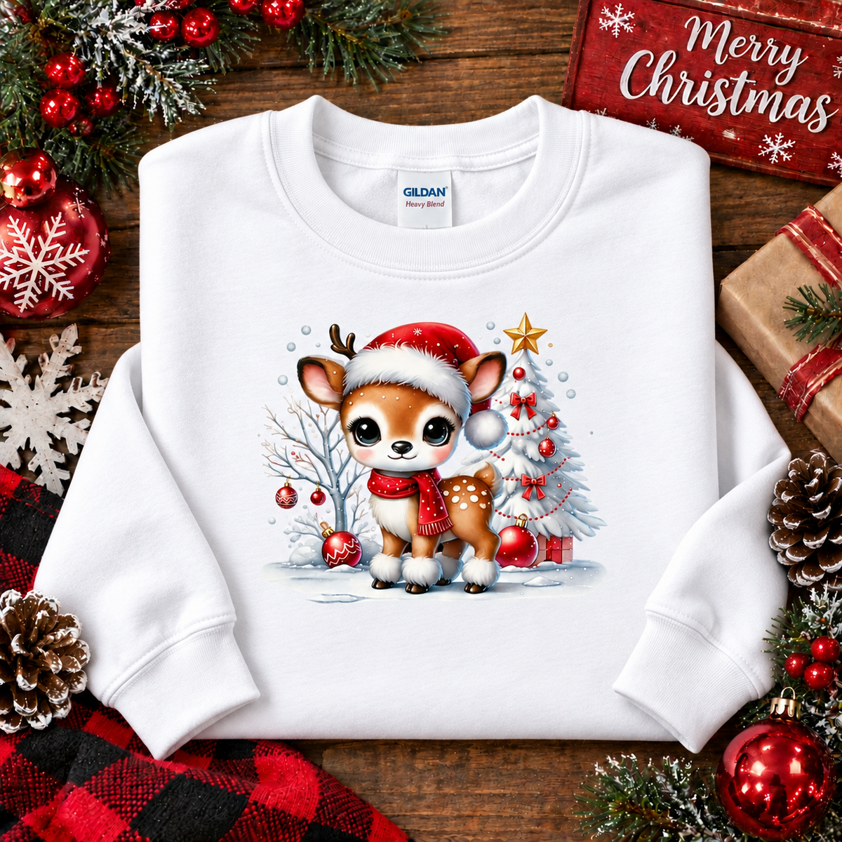 A white Gildan crewneck sweatshirt laid flat on a rustic wooden surface, decorated with Christmas ornaments, pinecones, evergreen branches, and a 'Merry Christmas' sign. The sweatshirt features a graphic of a cute cartoon fawn wearing a Santa hat and scarf, standing in front of a decorated Christmas tree.
