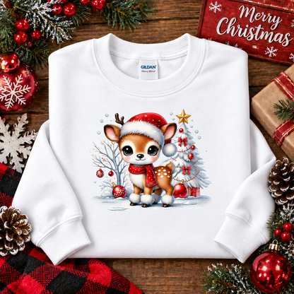 A white Gildan crewneck sweatshirt laid flat on a rustic wooden surface, decorated with Christmas ornaments, pinecones, evergreen branches, and a 'Merry Christmas' sign. The sweatshirt features a graphic of a cute cartoon fawn wearing a Santa hat and scarf, standing in front of a decorated Christmas tree.