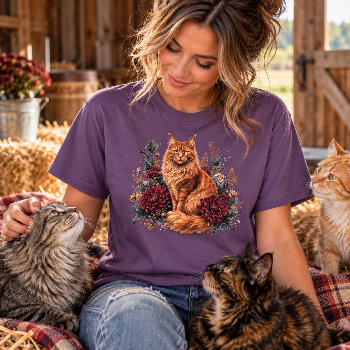 A woman wearing a heather purple graphic t-shirt with a detailed illustration of an orange tabby cat surrounded by pinecones and foliage, sitting in a rustic barn setting with three cats.