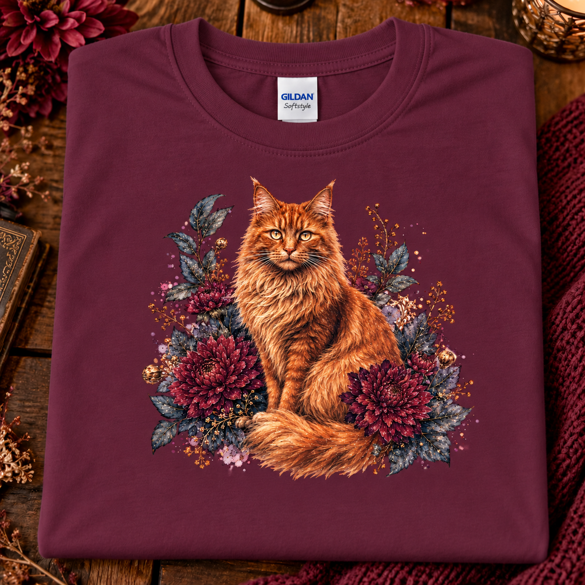 A folded maroon Gildan Softstyle t-shirt featuring a detailed graphic of an orange long-haired cat surrounded by dark red flowers and green leaves, displayed on a rustic wooden background with a knit blanket.