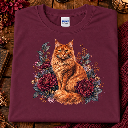 A folded maroon Gildan Softstyle t-shirt featuring a detailed graphic of an orange long-haired cat surrounded by dark red flowers and green leaves, displayed on a rustic wooden background with a knit blanket.