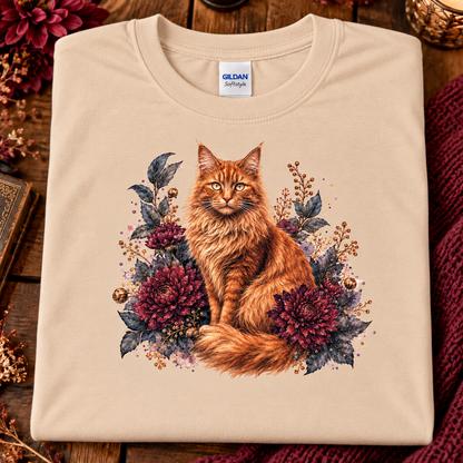 A natural colored Gildan t-shirt is folded and presented on a rustic wooden background with autumn-themed props like a book, dried flowers, and a knitted blanket. The t-shirt features a detailed graphic of a fluffy orange tabby cat surrounded by dark red flowers and green leaves.