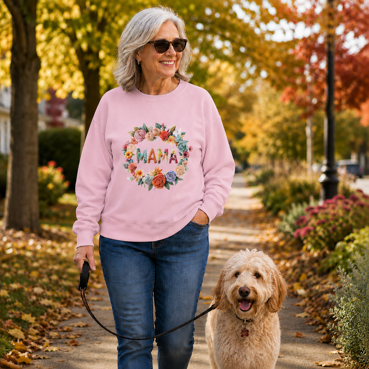 A woman walking a gold-colored dog on a leaf-covered path in autumn, wearing a light pink crewneck sweatshirt with a colorful floral wreath and the word 'MAMA' printed on the front.
